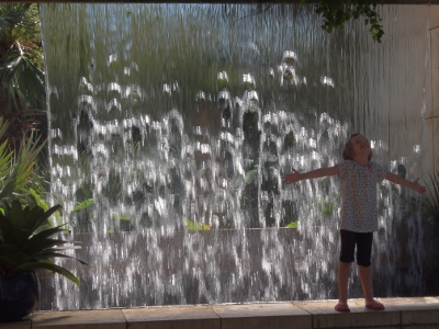 [An approximately 8 foot wide wall of water falling from a height of at least 6 feet. A young girl stands in front of it with her head tilted up and her arms outstretched to her sides. She is enjoying the spray of water coming from the fall.]